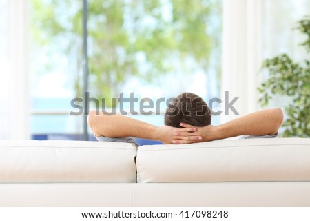 Rear view of a single man relaxed on a couch at home and looking the green background outdoors through the window in the livingroom