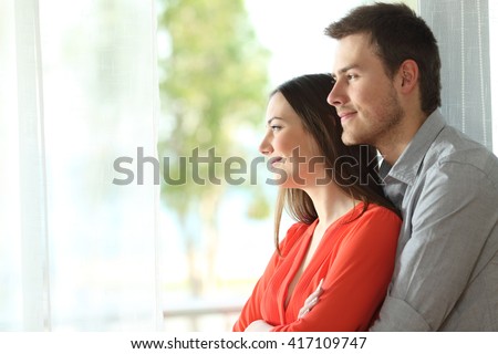 Side view portrait of a happy marriage standing hugging and looking outdoors through a window at home or hotel room with a green background