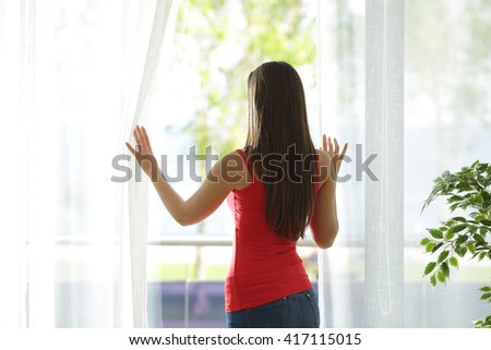Back view of a woman looking outdoors through a window and opening curtains at home