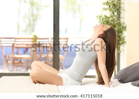 Side view of a woman breathing and sitting on a bed in an hotel room or home with a window in the background