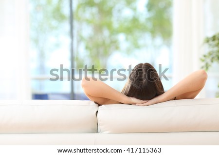 Rear view of a woman relaxing sitting on a couch with the hands on the head and looking outdoors through the window of the livingroom at home