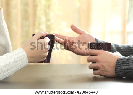 Back light profile of hands of 2 friends or couple talking holding coffee cups sitting in a table at home with a window in the background