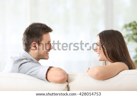 Side view of a couple or marriage talking sitting on a couch and looking each other at home with a white curtains in the background