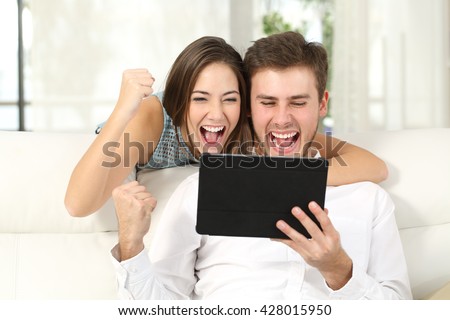 Front view of an excited couple winning online watching content in a tablet sitting on a couch in the living room at home