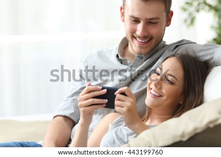 Happy couple enjoying media content in a smart phone sitting on a couch at home