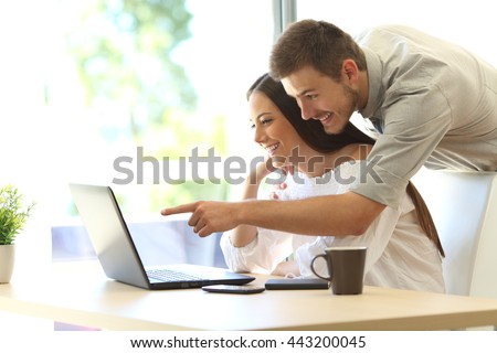 Side view of a happy couple searching information on line in a laptop on a table at home or hotel room with a window in the background