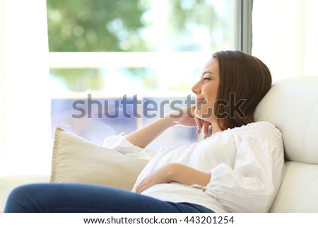 Pensive woman thinking sitting on a sofa and looking outdoors through a window with a green background