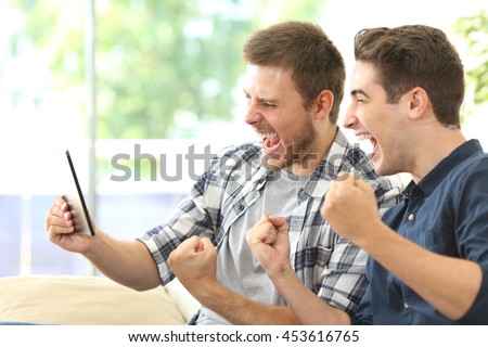 Two excited friends or roommates watching tv on line in a tablet sitting on a couch in the living room at home