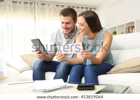 Happy couple checking bank account on line in a tablet sitting on a couch in the living room at home