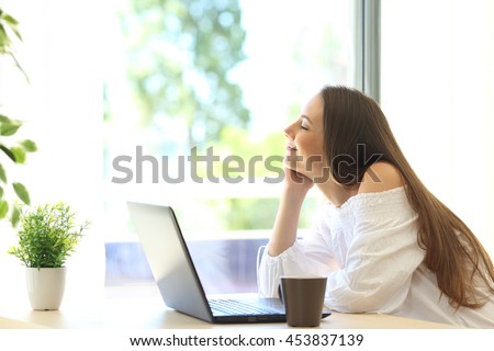 Side view of a happy girl with a laptop relaxing sitting near a window of a house interior