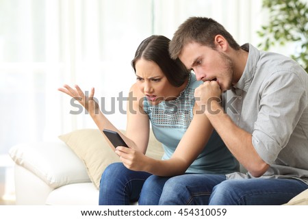 Angry couple with mobile phone sitting on a couch in the living room at home