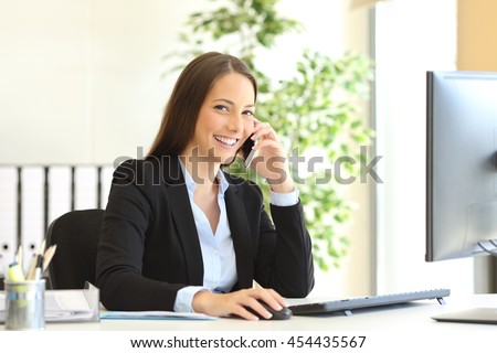 Portrait of a happy executive wearing suit calling customer service on the phone and looking at camera sitting on a desk in the office