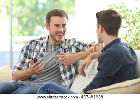 Two friends talking sitting in a couch in the living room with a window in the background at home