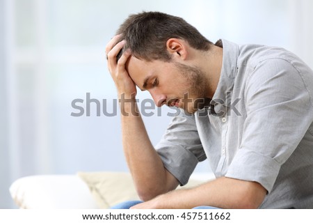 Side view of a sad man with a hand on the head sitting on a couch in the living room at home