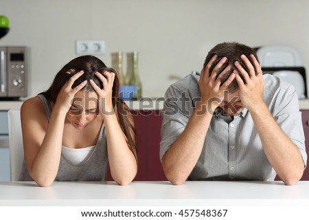 Front view of a sad couple with hands on head sitting in the kitchen of a house