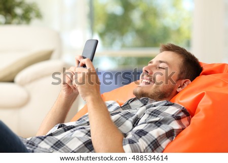 Relaxed man using a smart phone lying on an orange pouf in the living room at home with a window in the background