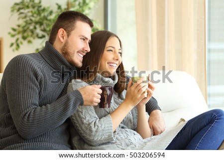 Affectionate couple wearing sweater holding hot coffee cups sitting on a sofa in the living room at home in winter