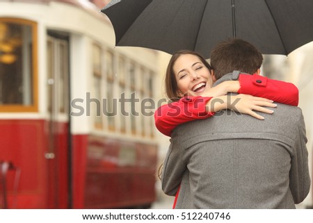 Encounter after a travel of a happy couple hugging in the street in a tram station in a rainy day under an umbrella