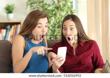 Two amazed roommates watching offers on line with a smart phone sitting on a couch in the living room of a house