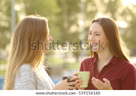 Two happy friends talking holding tea mug outdoors in a park with a green background at sunset with a warm back light