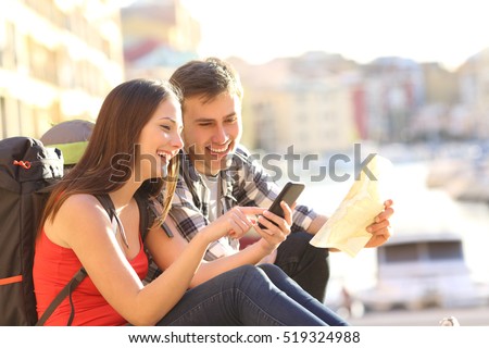 Two tourists searching location on a smart phone sitting on the floor of a travel destination in the coast with a warm light in the background