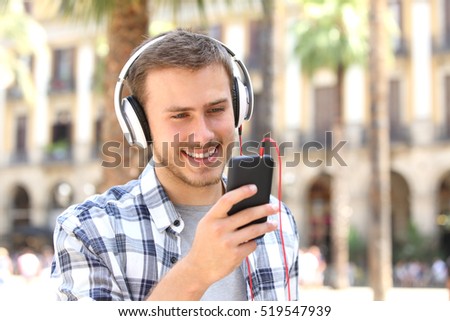 Front view of a happy guy with headphones listening music on line and watching media in a smartphone in the street
