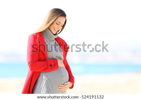 Happy pregnant woman wearing a red jacket looking her belly on the beach in winter