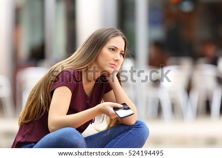 Sad girl waiting for a mobile phone call or message from her boyfriend sitting in a bench outside in the street with an urban background