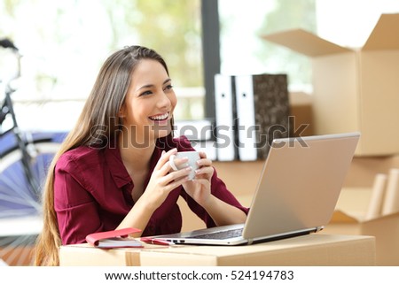 Entrepreneur moving office working on line with a laptop and thinking holding a coffee cup with her belongings in carton boxes in the background