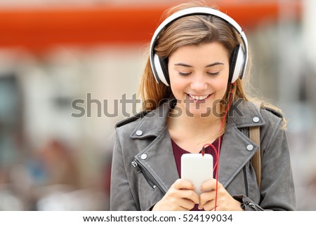 Front view of a fashion girl listening music with headphones and smart phone on line and walking towards camera in the street