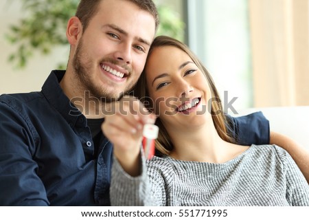 Happy couple of owners showing house keys to the camera sitting on a sofa in the living room at home