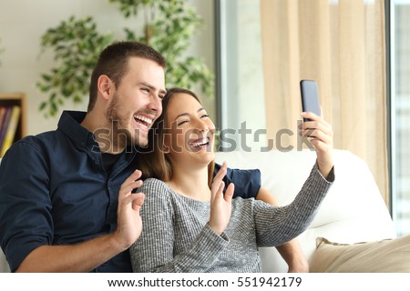 Happy casual couple greeting in a phone video call on line sitting on a sofa in the living room at home with a window in the background