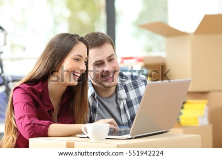 Happy couple searching online with a laptop over a cardboard box sitting on the floor while moving apartment