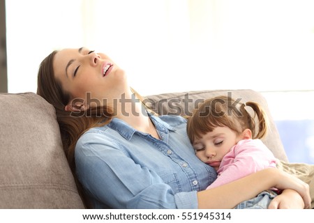 Tired mother sleeping embracing to her asleep baby daughter sitting on a sofa at home