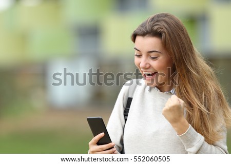 Excited student reading good news on line in a smart phone in the street with the university building in the background