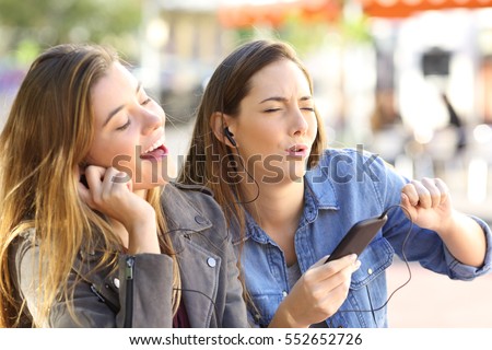 Joyful friends listening to music on line from a smart phone and singing sharing earphones in the street
