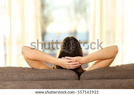 Back view of a single woman relaxing looking outside through a window sitting on a sofa in the living room at home with a warm light