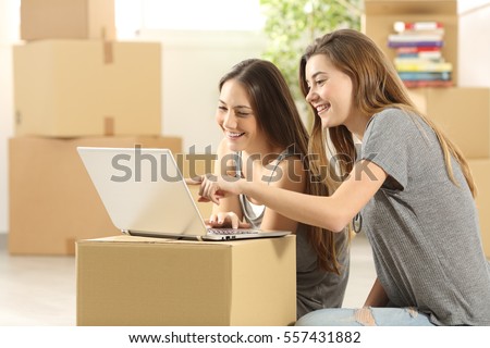 Happy roommates searching on line with a laptop on a cardboard box and moving home sitting on the floor of the living room