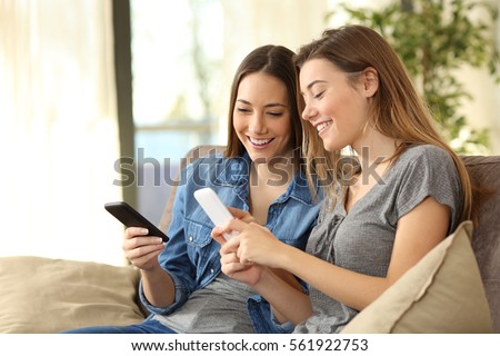 Two roommates using their smart phones on line at home sitting on a sofa in the living room at home