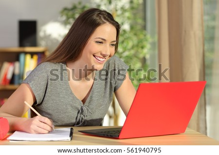 Student taking notes and learning on line with a red laptop sitting in a table in the living room at home