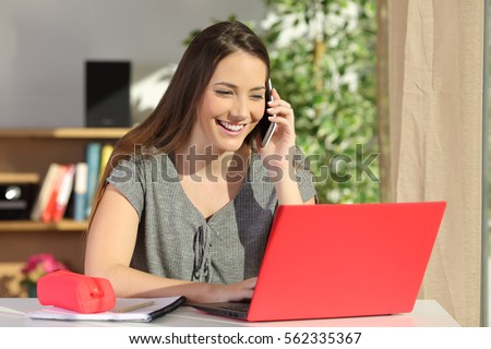 Happy student asking information by phone consulting a laptop on line sitting in a table at home with a colorful background