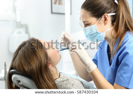 Dentist wearing mask gloves and blue uniform examining a patient teeth with a dental probe and a mirror in a box with medical equipment in the background