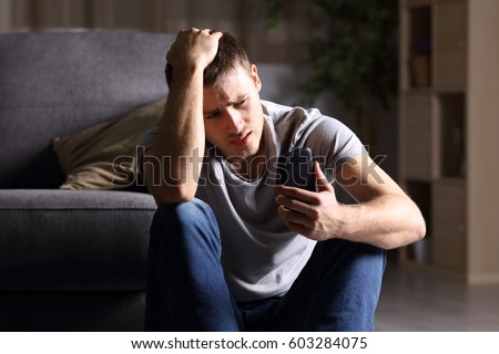 Single sad man checking mobile phone sitting on the floor in the living room at home with a dark background