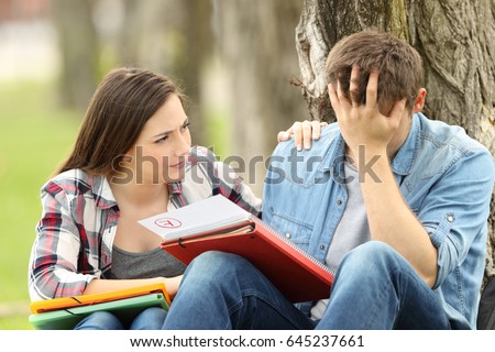 Friend comforting to a sad student with failed exam sitting on the grass in a park