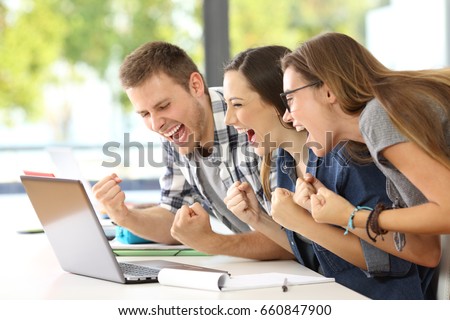 Side view of three excited students reading good news together on line in a laptop sitting in a desk in a classroom