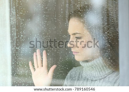 Portrait of a sad girl looking through a window in a rainy day at home