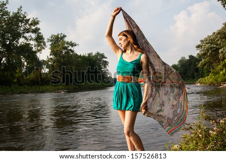 Young woman with a teal green dress stands near a river and lake holding teal scarf in the wind.   She is relaxed and serene while feeling the summer breeze.