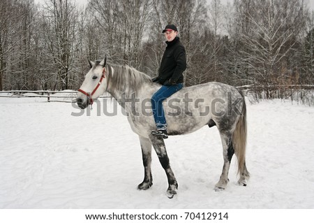 The young guy in a black jacket and a cap sits on is light grey horse on a winter wood glade
