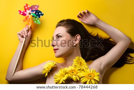 Topless portrait of a beautiful young woman covered with yellow flowers holding colorful pin wheel isolated on yellow background