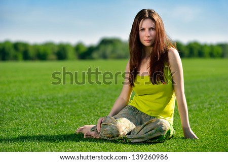 Portrait of young beautiful brunette woman with long hair wearing bright dress, sitting on green grass at summer meadow.